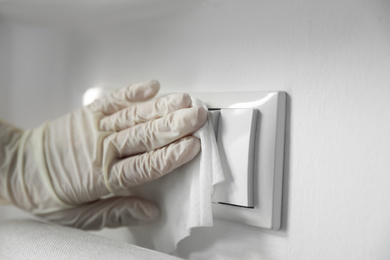 Photo of Person in latex gloves cleaning light switch with wet wipe indoors, closeup