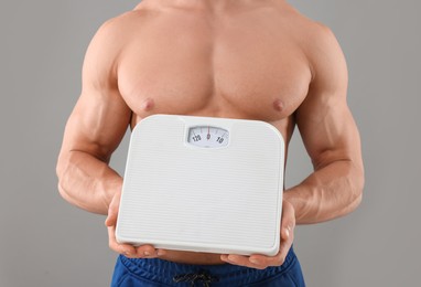 Athletic man holding scales on grey background, closeup. Weight loss concept