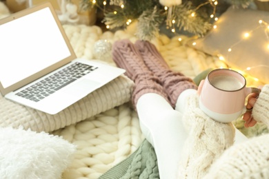 Woman with cup of coffee and laptop sitting near Christmas tree, closeup