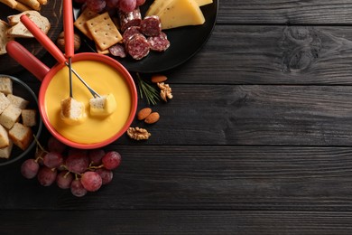 Photo of Pot of tasty cheese fondue and snacks on black wooden table, flat lay. Space for text