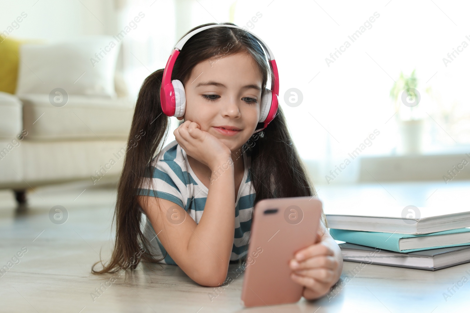 Photo of Cute little girl with headphones and smartphone listening to audiobook at home