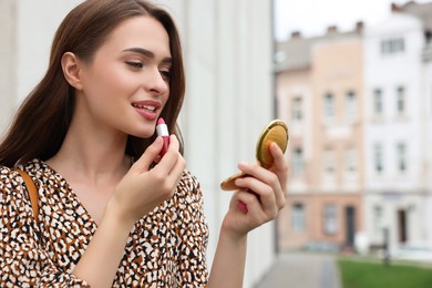 Beautiful young woman with cosmetic pocket mirror applying lipstick outdoors, space for text