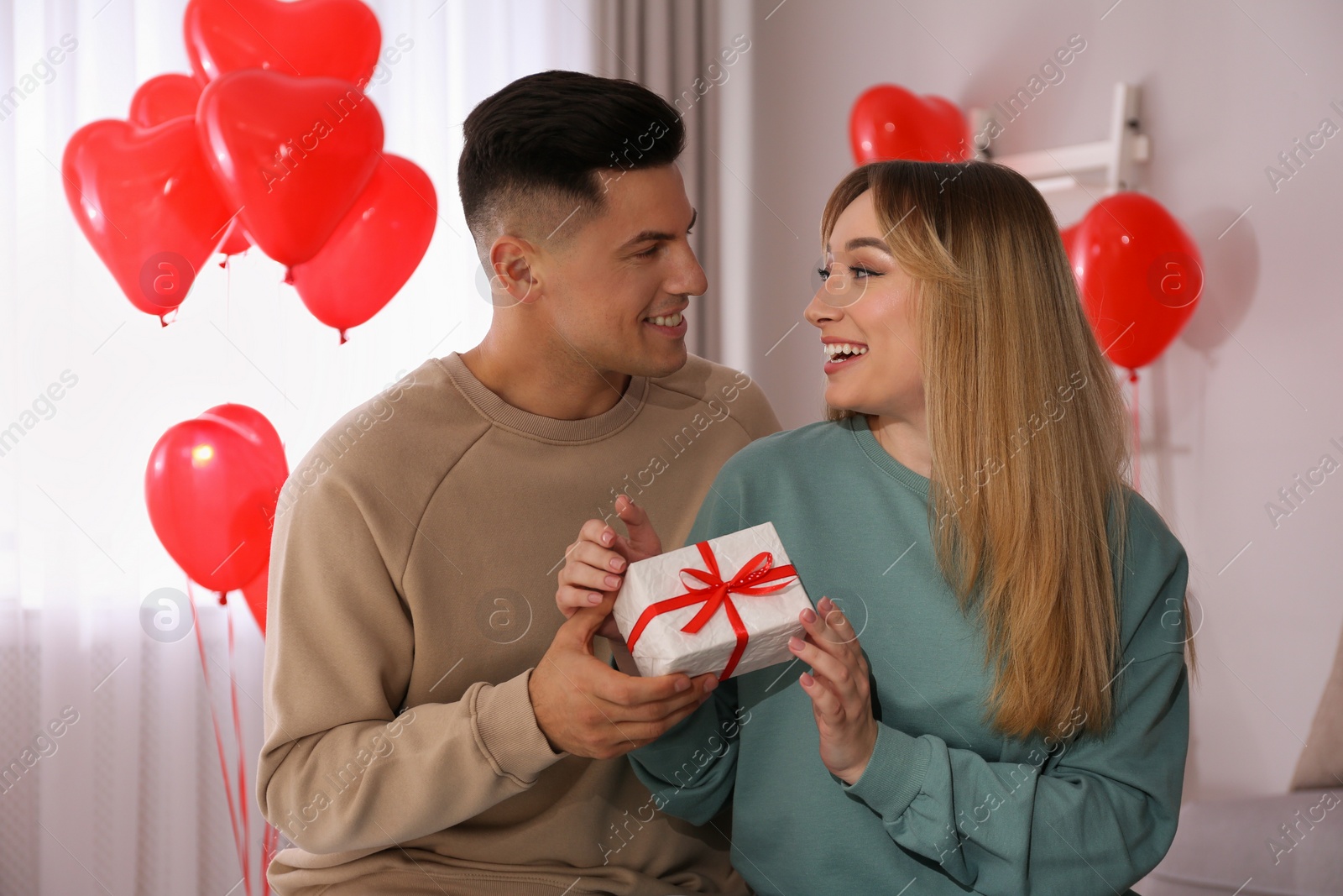 Photo of Man presenting gift to his girlfriend in room decorated with heart shaped balloons. Valentine's day celebration