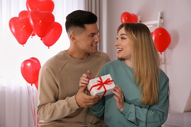 Man presenting gift to his girlfriend in room decorated with heart shaped balloons. Valentine's day celebration