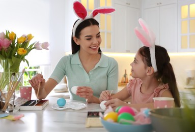 Happy mother with her cute daughter painting Easter eggs at table in kitchen