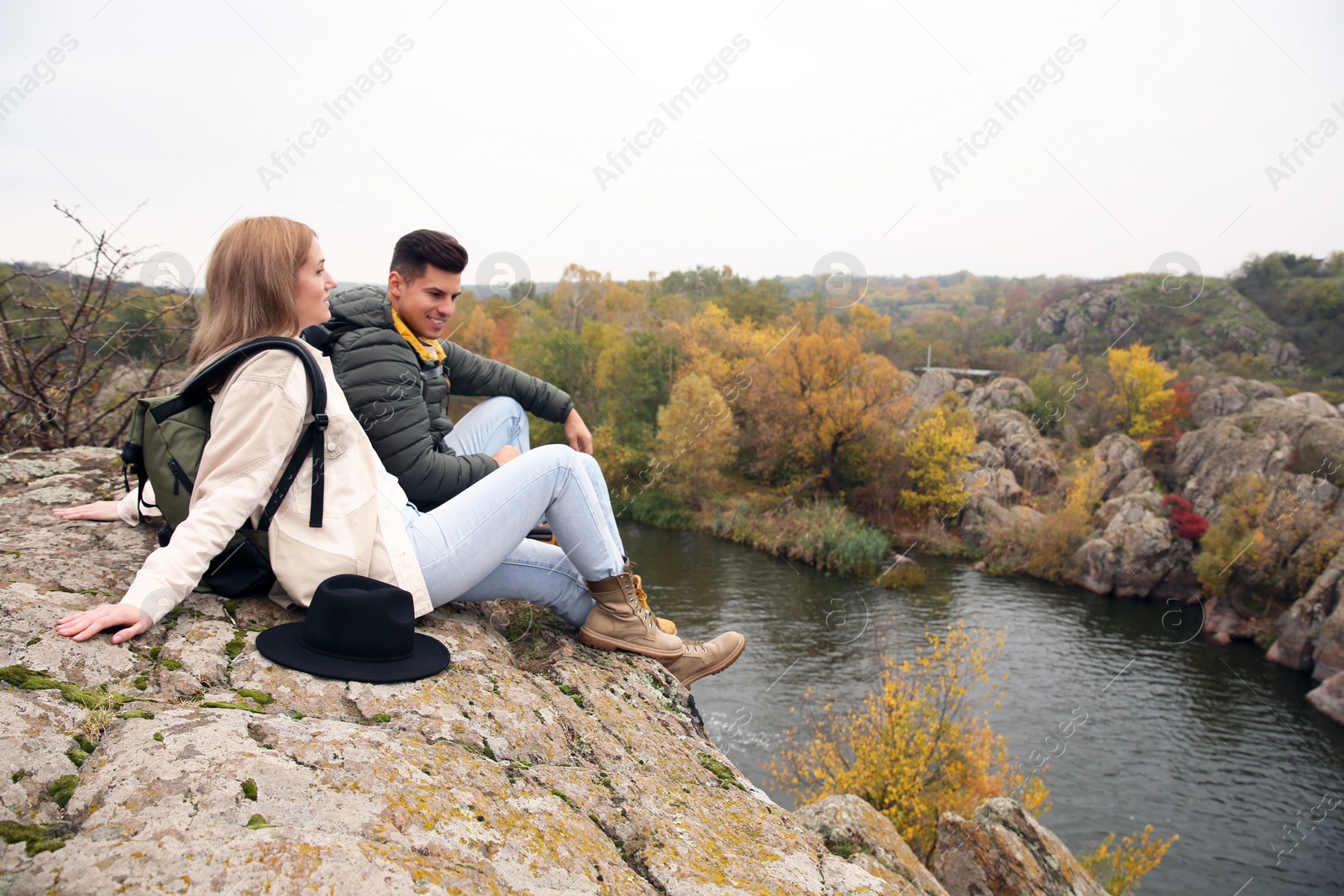 Photo of Couple of hikers with travel backpacks sitting on steep cliff near mountain river