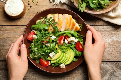 Woman holding bowl of fresh salad with arugula and chicken at wooden table, top view