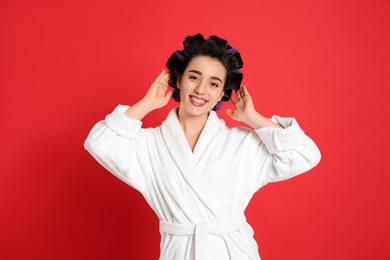 Photo of Happy young woman in bathrobe with hair curlers on red background