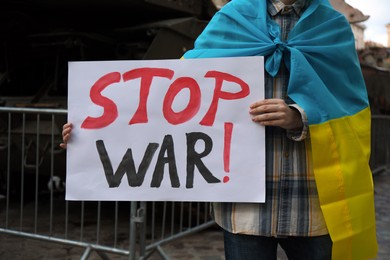 Photo of Woman wrapped in Ukrainian flag holding poster with words Stop War outdoors, closeup