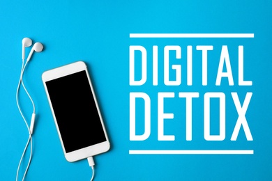Image of Text Digital Detox, modern phone with earphones on light blue background, top view.