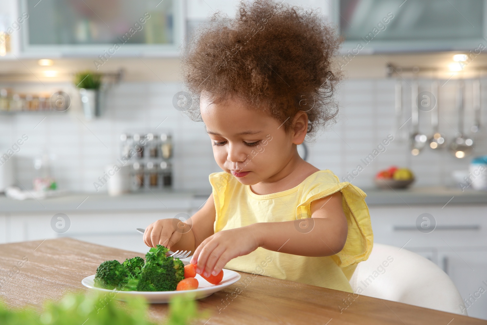 Photo of Cute African-American girl eating vegetables at table in kitchen