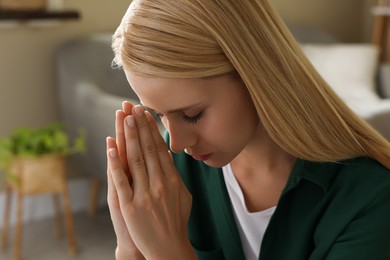 Religious young woman with clasped hands praying indoors