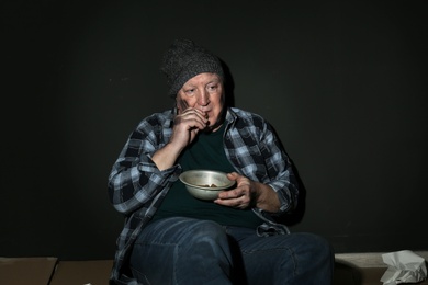 Poor senior man with bread and bowl on floor near dark wall