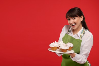 Photo of Happy professional confectioner in apron holding delicious cupcakes on red background. Space for text