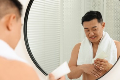 Photo of Handsome man applying body cream onto his hand near mirror indoors