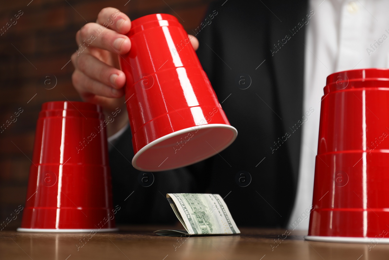 Photo of Shell game. Man showing dollar banknote under cup at wooden table, closeup