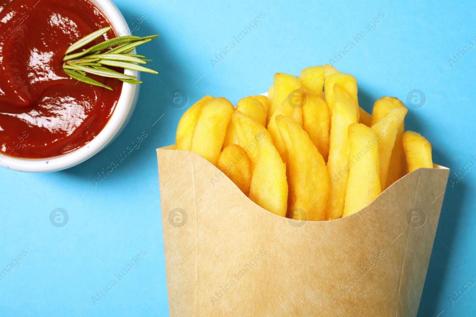 Photo of Paper cup with French fries, rosemary and ketchup on light blue table, flat lay