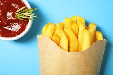 Paper cup with French fries, rosemary and ketchup on light blue table, flat lay