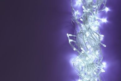 Photo of Glowing Christmas lights on dark violet background, top view. Space for text