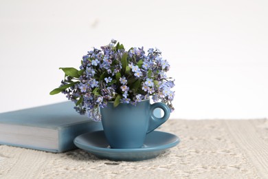 Beautiful forget-me-not flowers in cup, book and crochet tablecloth on table against white background, closeup