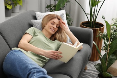 Photo of Woman reading book on sofa surrounded by beautiful potted houseplants at home