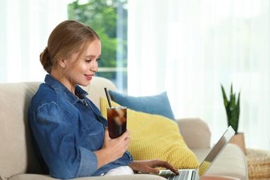 Photo of Young woman with glass of cola working on laptop at home. Refreshing drink