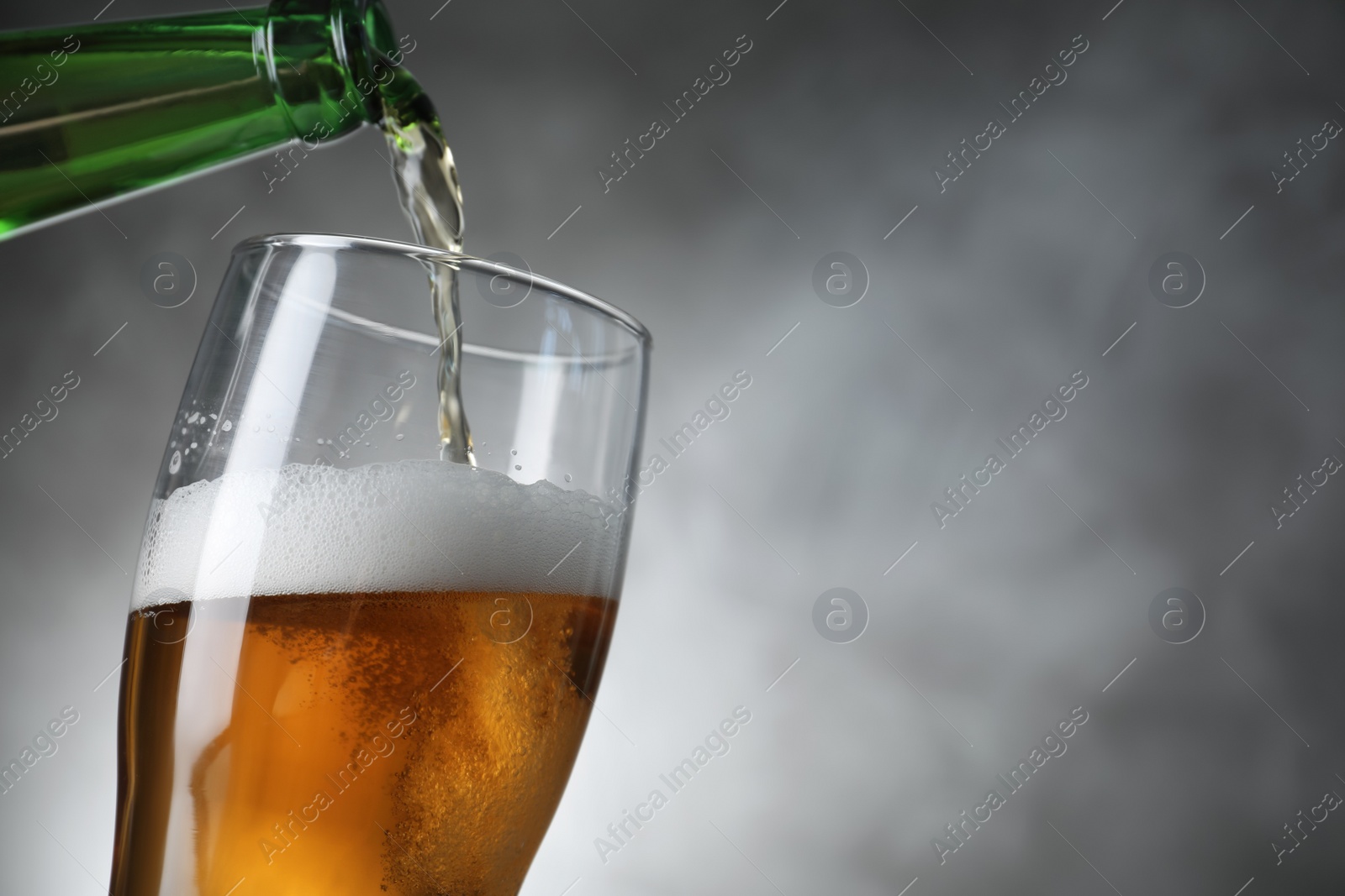 Photo of Pouring cold tasty beer from bottle into glass on grey background, closeup. Space for text