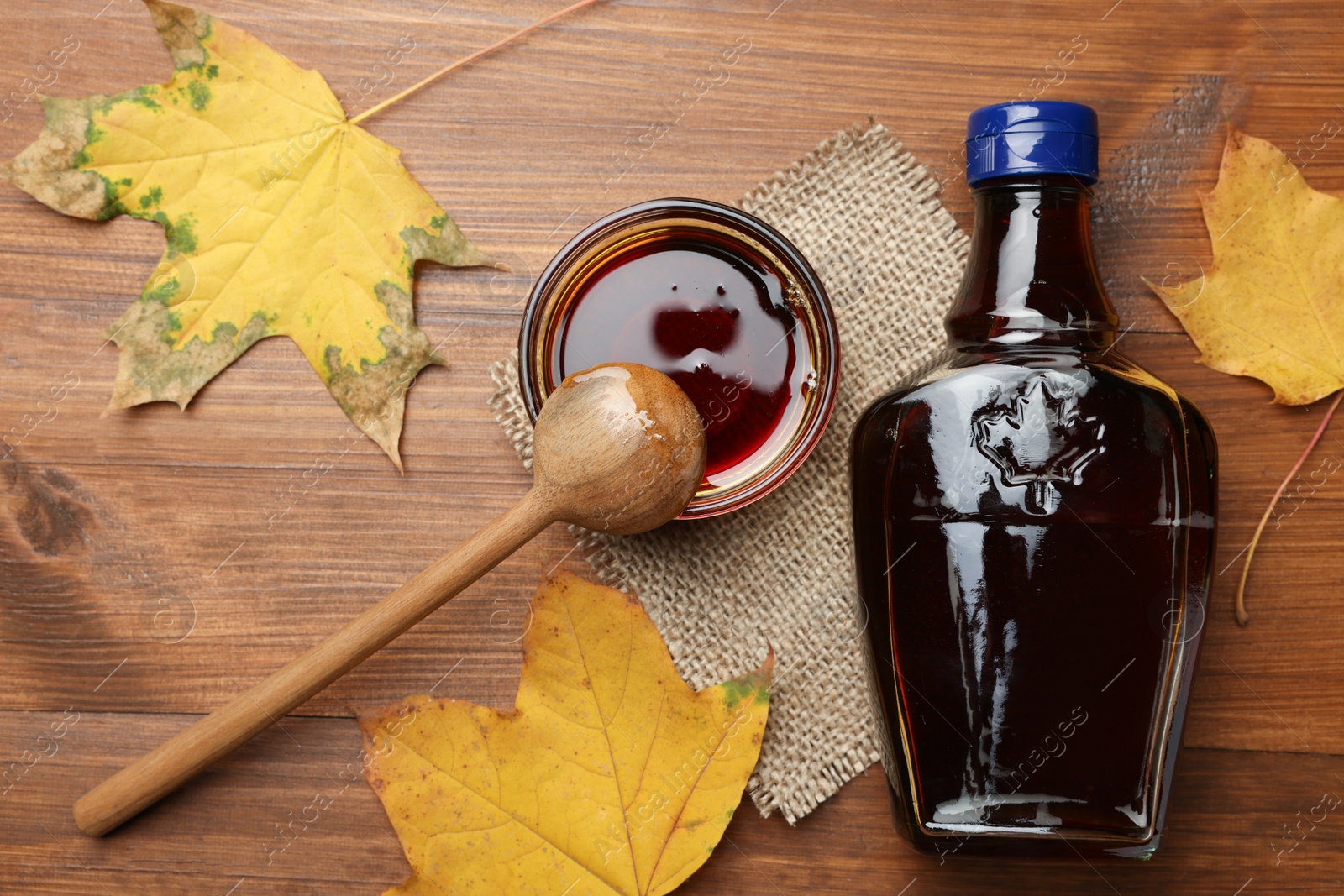 Photo of Bottle of tasty maple syrup, bowl and dry leaves on wooden table, flat lay