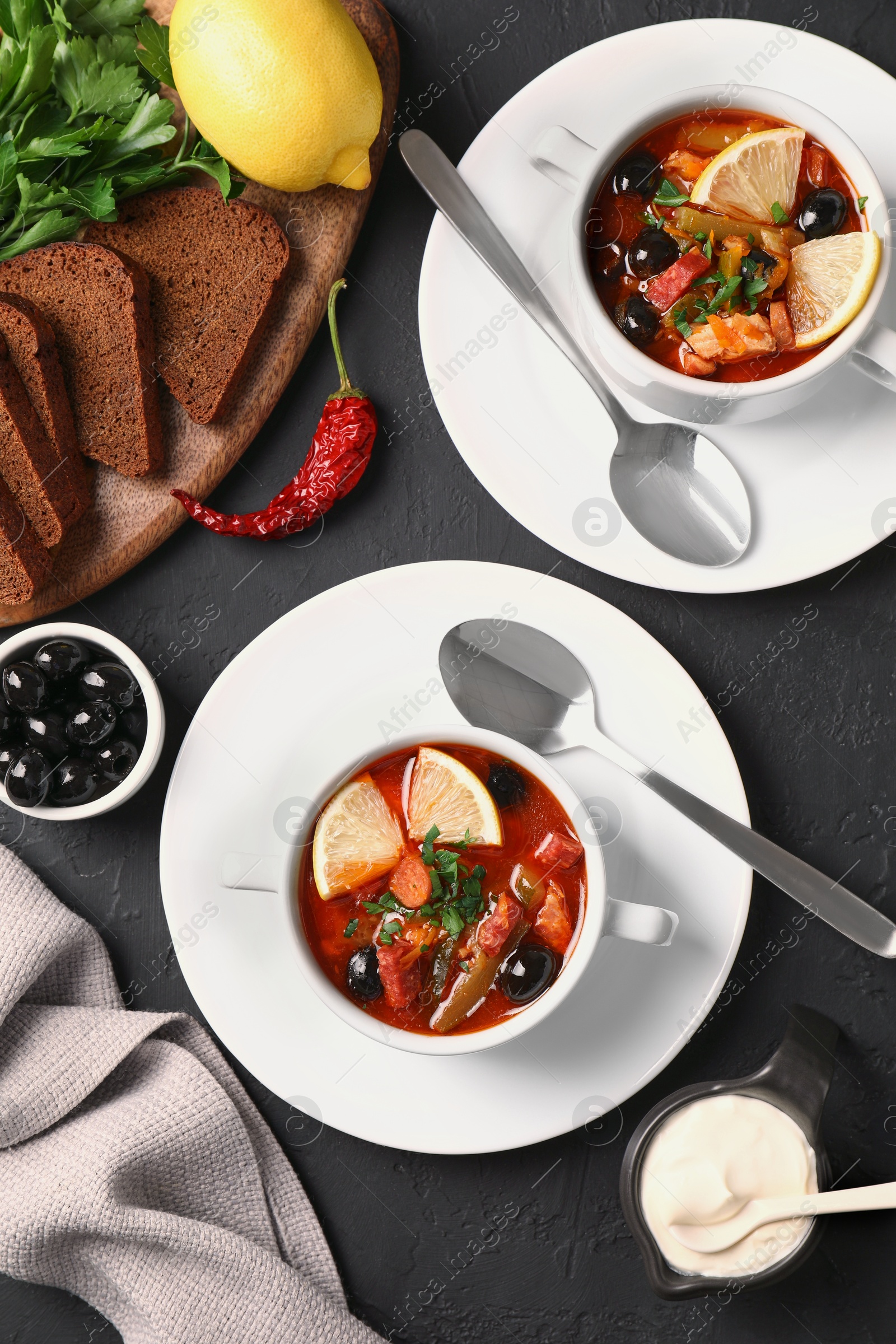 Photo of Meat solyanka soup with sausages, olives and vegetables served on dark grey textured table, flat lay