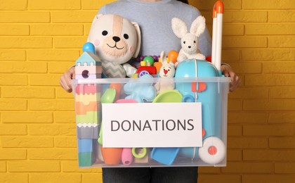 Photo of Woman holding donation box full of different toys near yellow brick wall, closeup