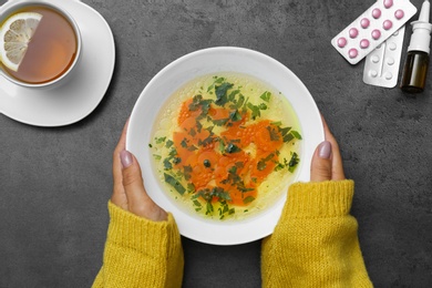 Photo of Woman with bowl of soup at grey table, top view. Flu treatment