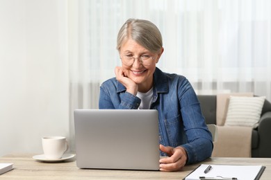Photo of Beautiful senior woman using laptop at wooden table indoors