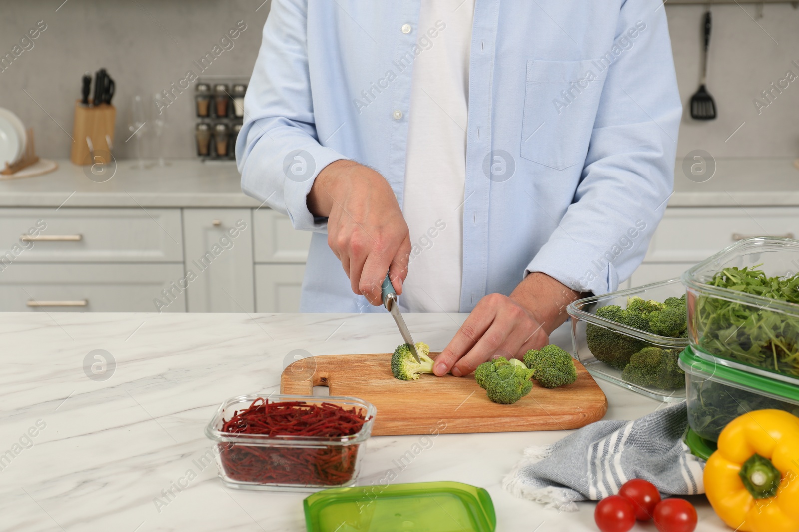 Photo of Man cutting fresh broccoli with knife near containers at white marble table in kitchen, closeup and space for text. Food storage