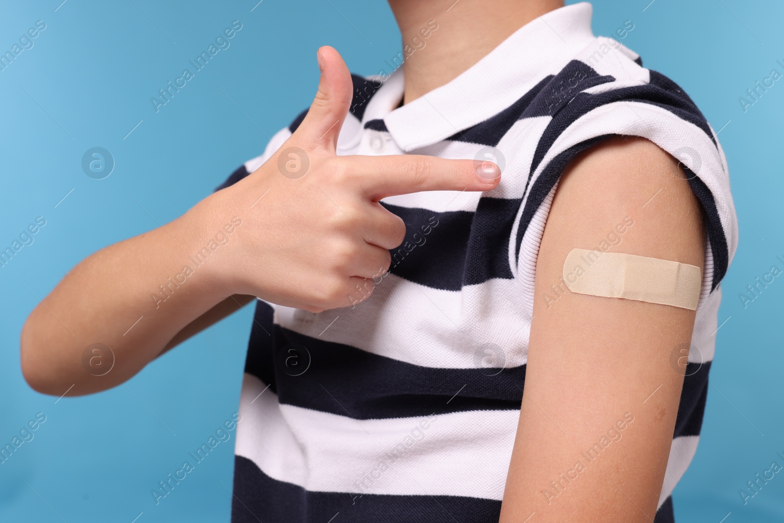 Photo of Boy pointing at sticking plaster after vaccination on his arm against light blue background, closeup