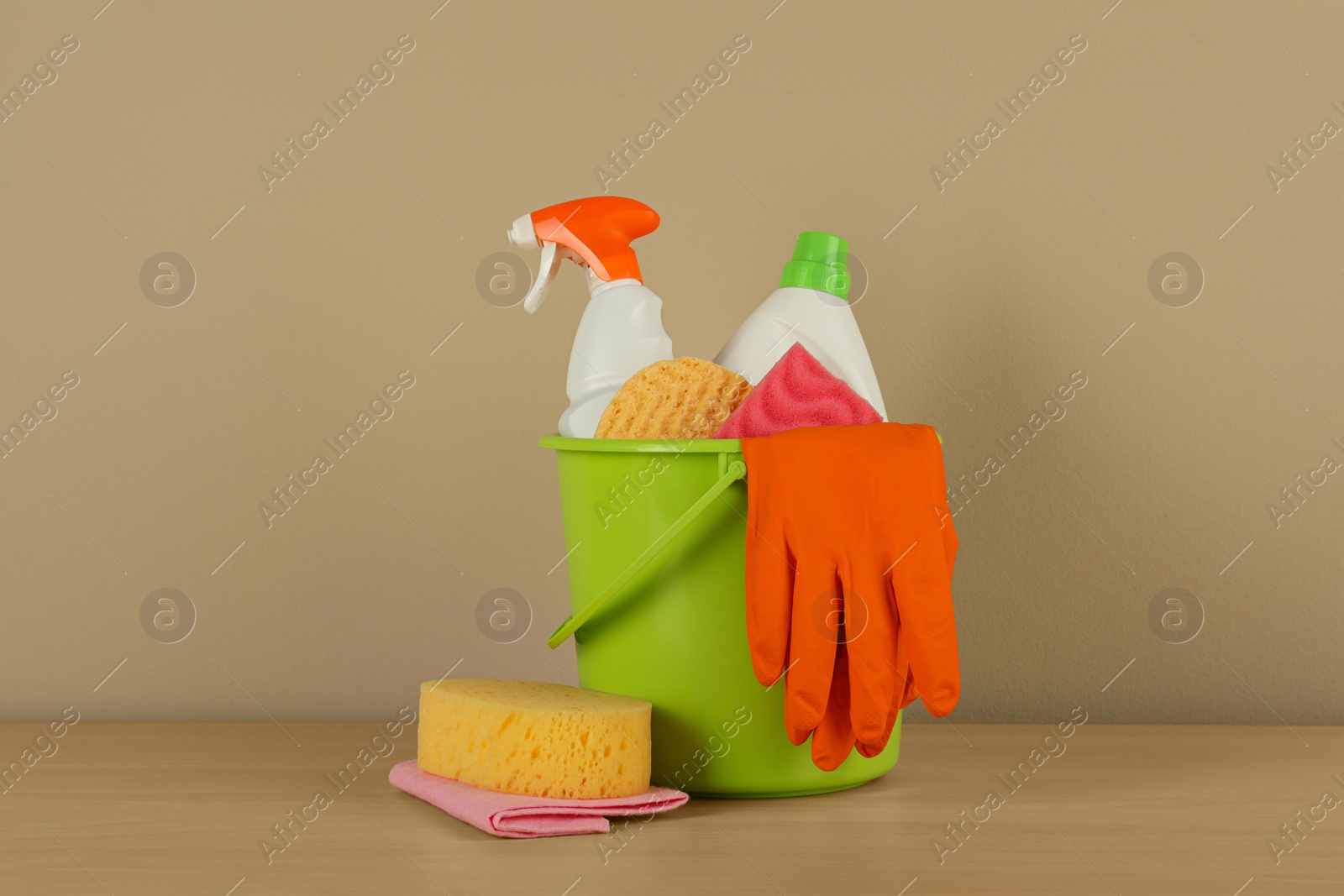 Photo of Bucket with different cleaning supplies on wooden floor near beige wall