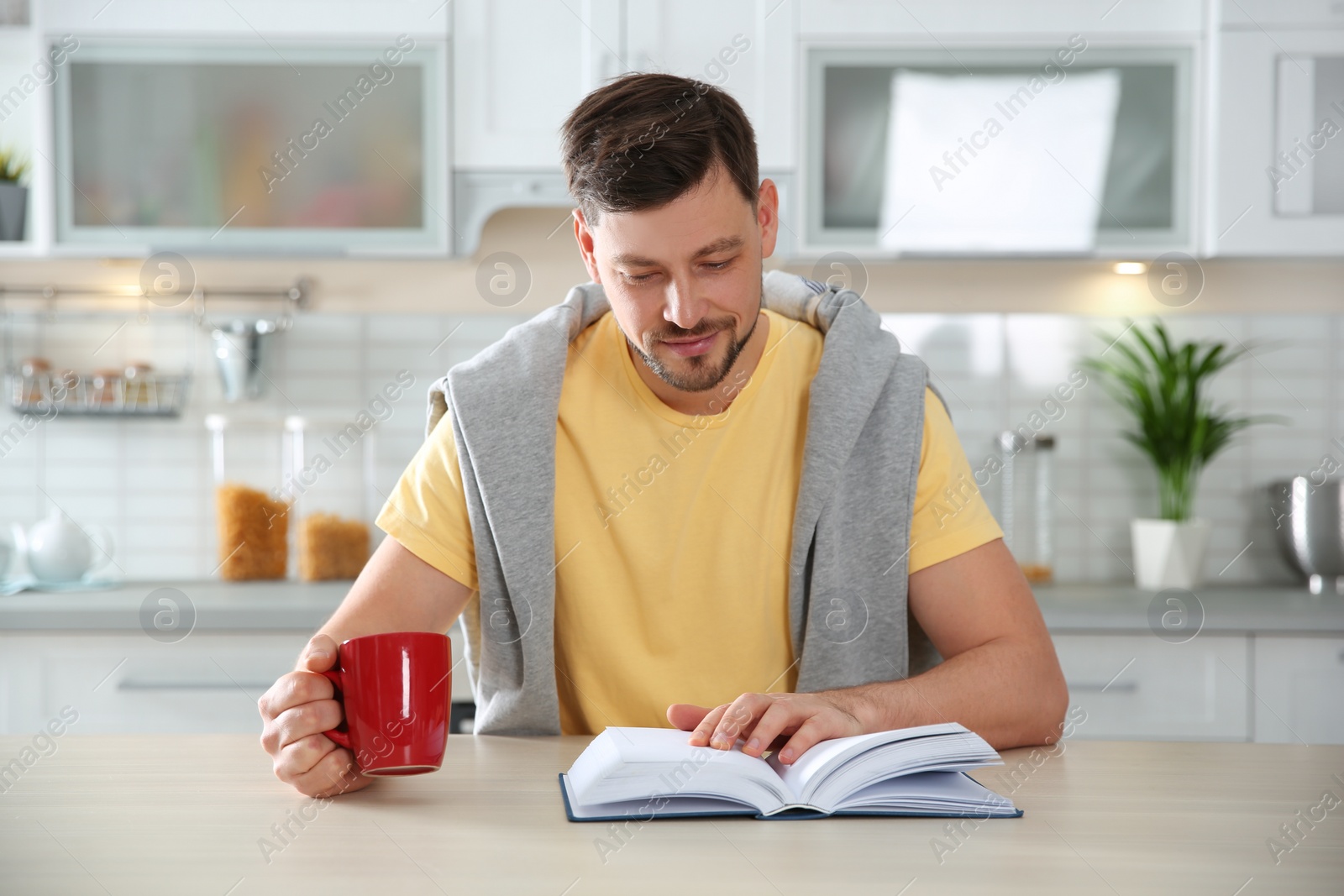 Photo of Handsome man with cup of coffee reading book at table in kitchen