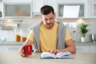 Handsome man with cup of coffee reading book at table in kitchen