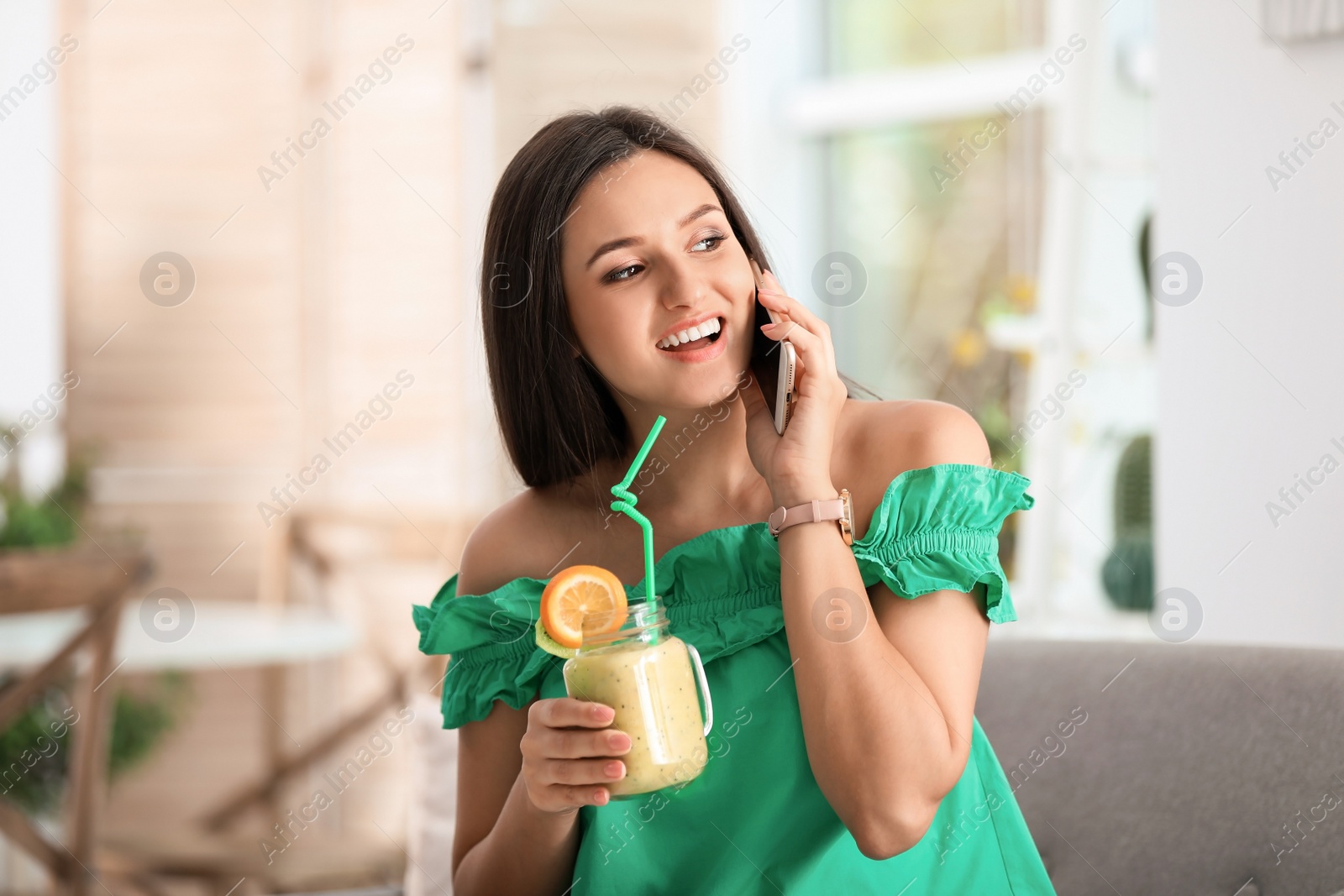 Photo of Young woman using mobile phone while drinking tasty healthy smoothie at table, indoors