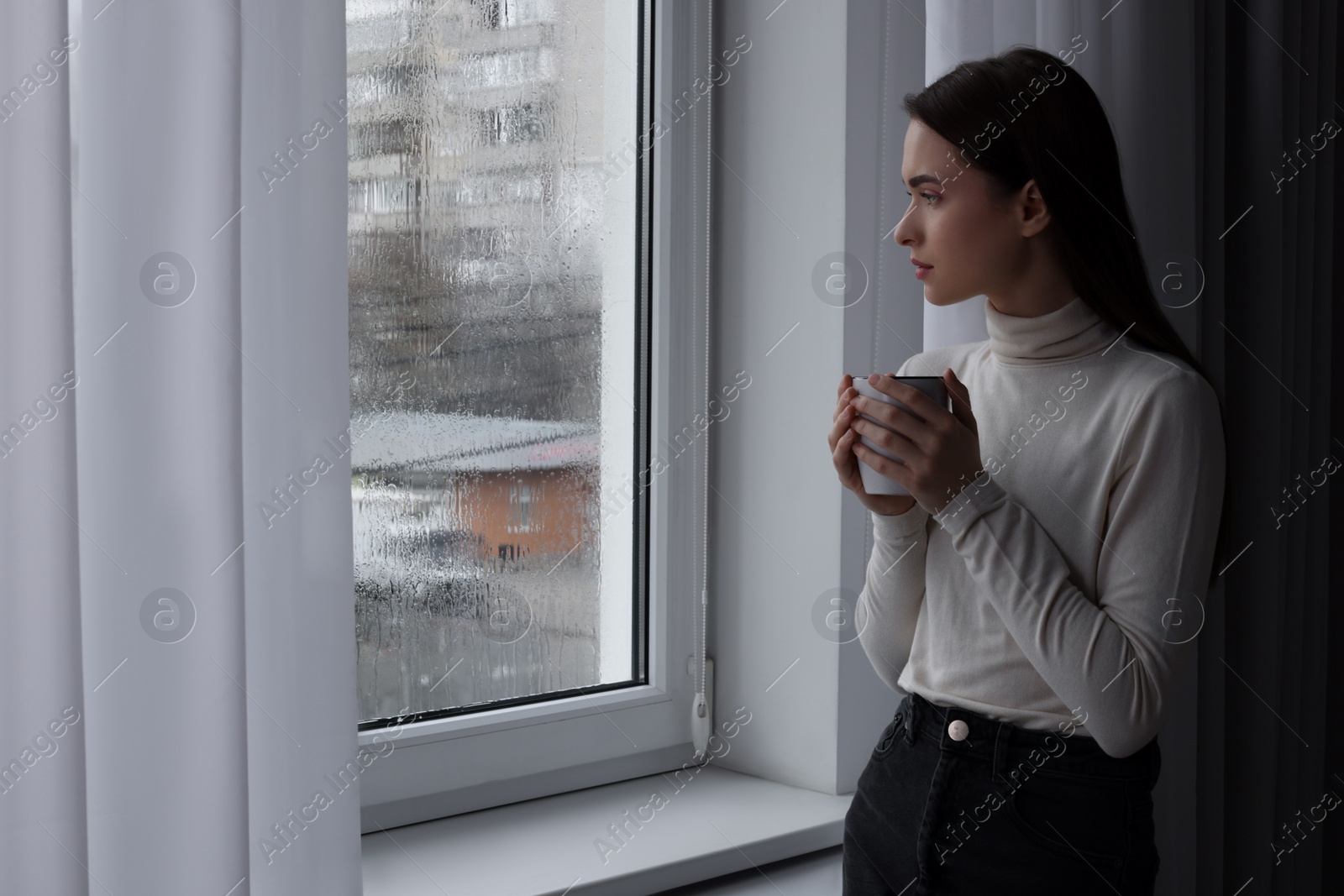 Photo of Melancholic young woman with drink looking out of window on rainy day, space for text. Loneliness concept