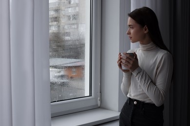 Photo of Melancholic young woman with drink looking out of window on rainy day, space for text. Loneliness concept