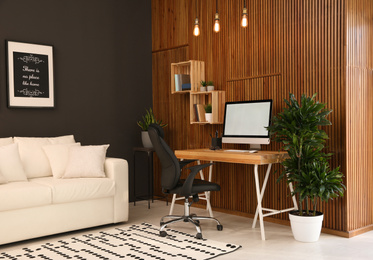 Comfortable workplace with computer near wooden wall in stylish room interior. Home office design