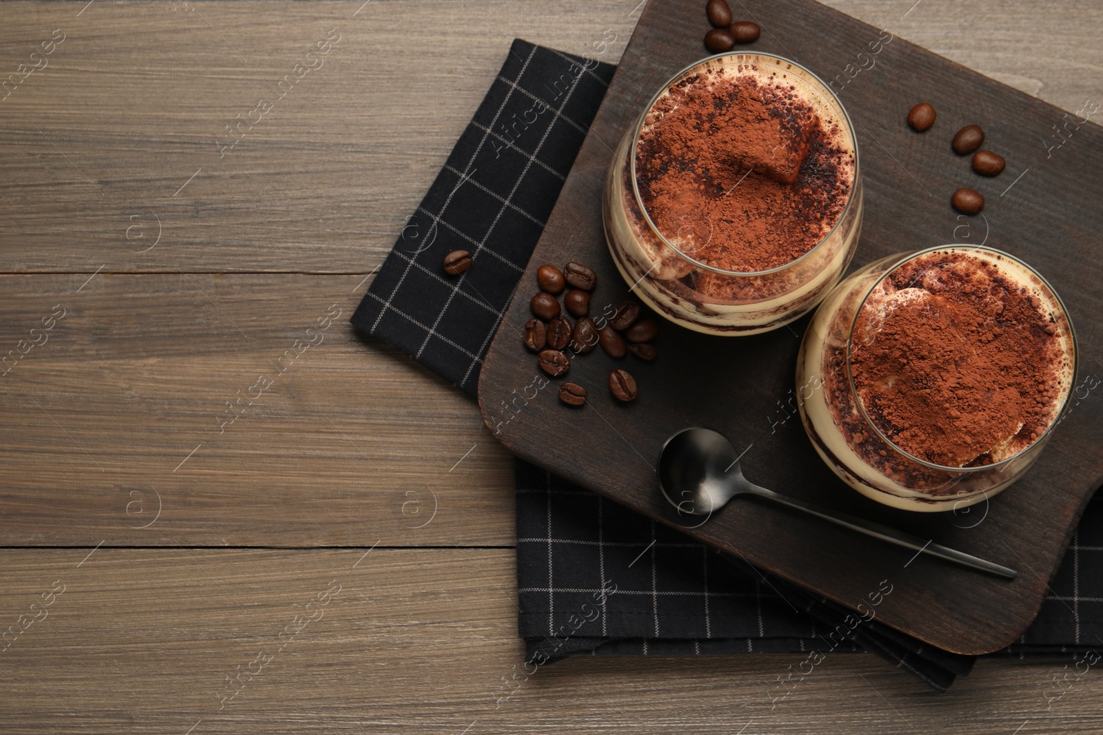 Photo of Delicious tiramisu in glasses, spoon and coffee beans on wooden table, top view. Space for text