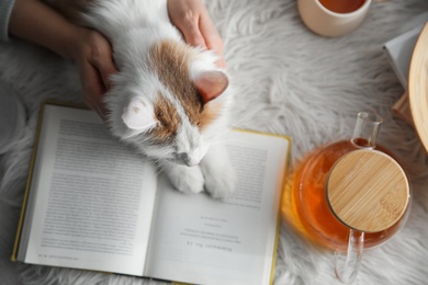 Photo of Woman with cute fluffy cat, tea and book on faux fur, top view