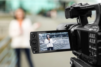 Photo of Young female journalist with microphone working on city street, focus on camera display