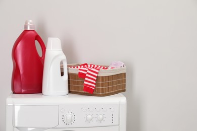 Baby clothes in wicker basket and laundry detergents on washing machine near light wall, space for text