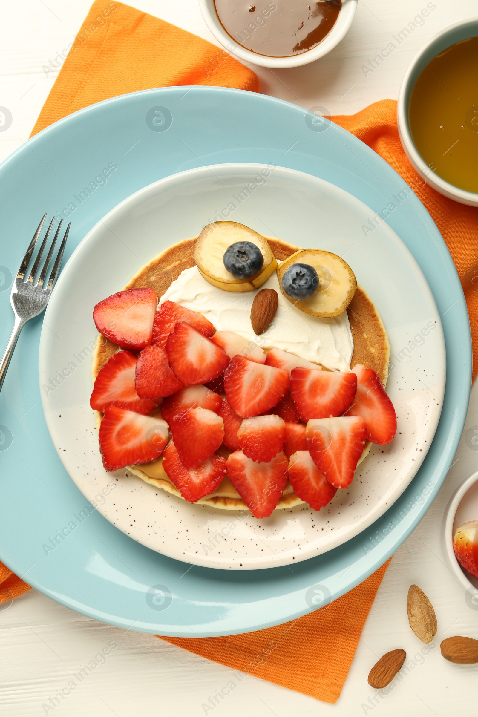 Photo of Creative serving for kids. Plate with cute owl made of pancakes, strawberries, cream, banana and almond on white wooden table, flat lay