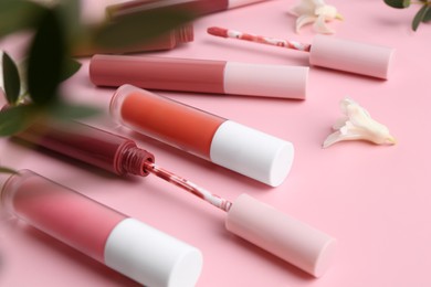 Photo of Different lip glosses, applicators, flowers and green leaves on pink background, closeup