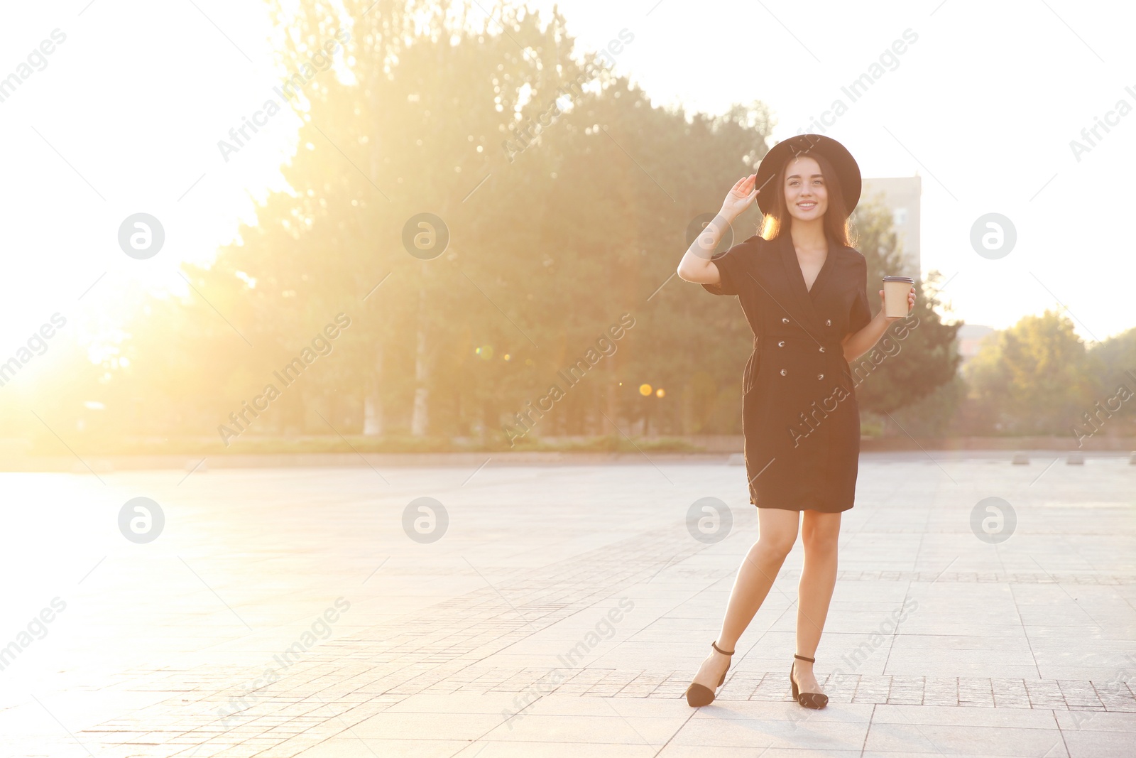 Photo of Beautiful young woman in stylish black dress and hat with cup of coffee on city street