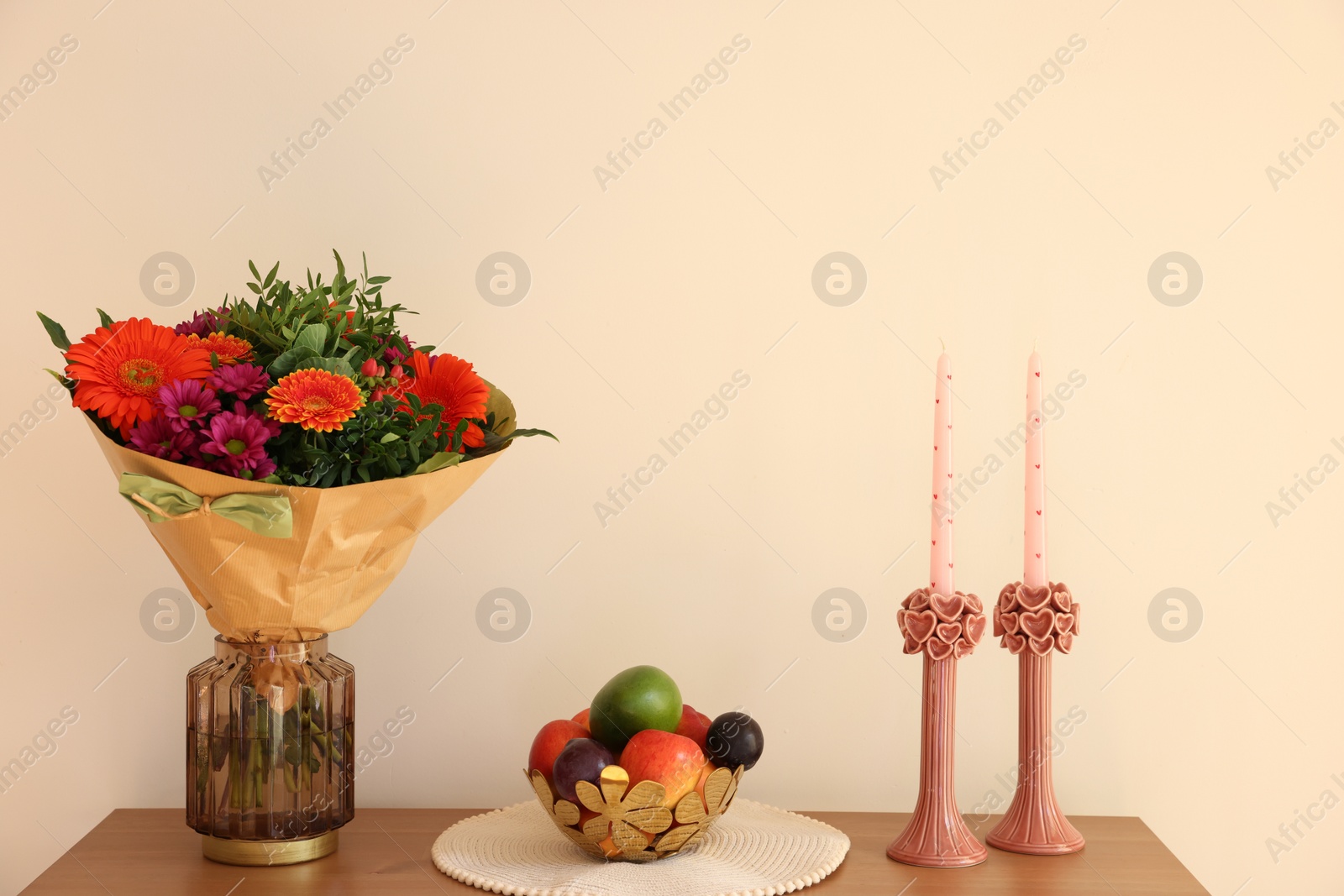 Photo of Bouquet of flowers, bowl with fresh fruits and candles on wooden table near white wall