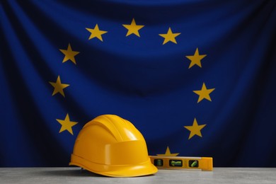 Photo of Yellow hard hat and building level on light grey table against flag of European Union